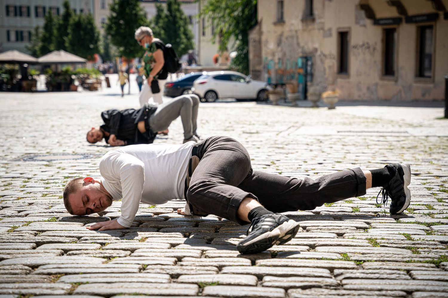 Two men lying on cobblestone plaza performing contemporary dance in bright sunlight.