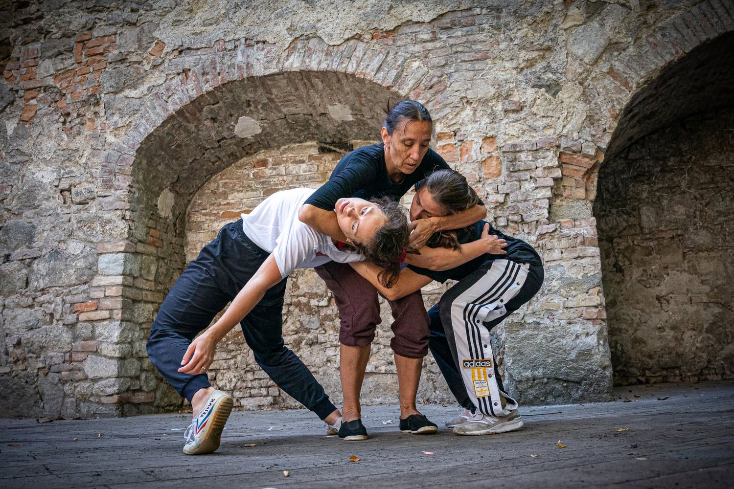 Three dancers performing an intimate contemporary dance hold outdoors against a rustic stone wall.