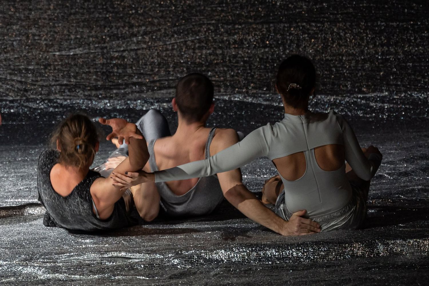 Three dancers in gray costumes sit closely together on a rain-soaked reflective stage floor.