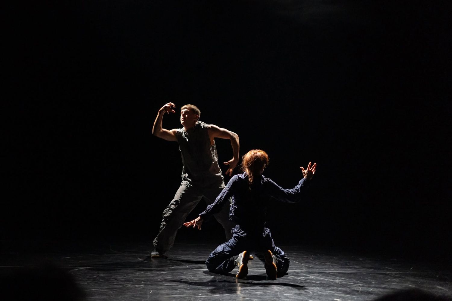 Two contemporary dancers perform an intense duet on a dark stage, one standing and one crouching low.