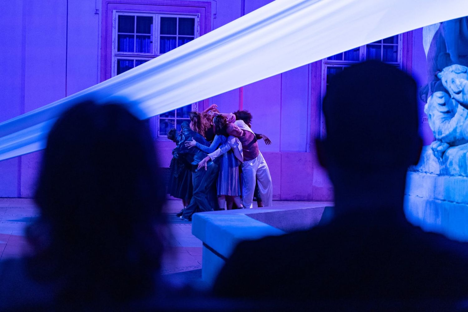 Performers on a dramatic blue-lit outdoor stage with flowing white fabric, watched by silhouetted audience.
