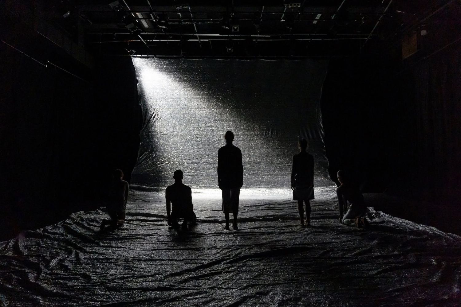 Silhouetted performers stand and kneel dramatically under a single spotlight on a dark tarp-covered stage.