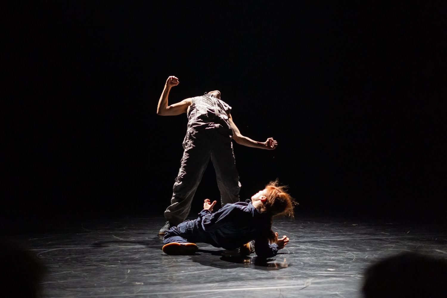 Two modern dancers perform a dramatic floor-level contemporary dance piece on a dark stage.