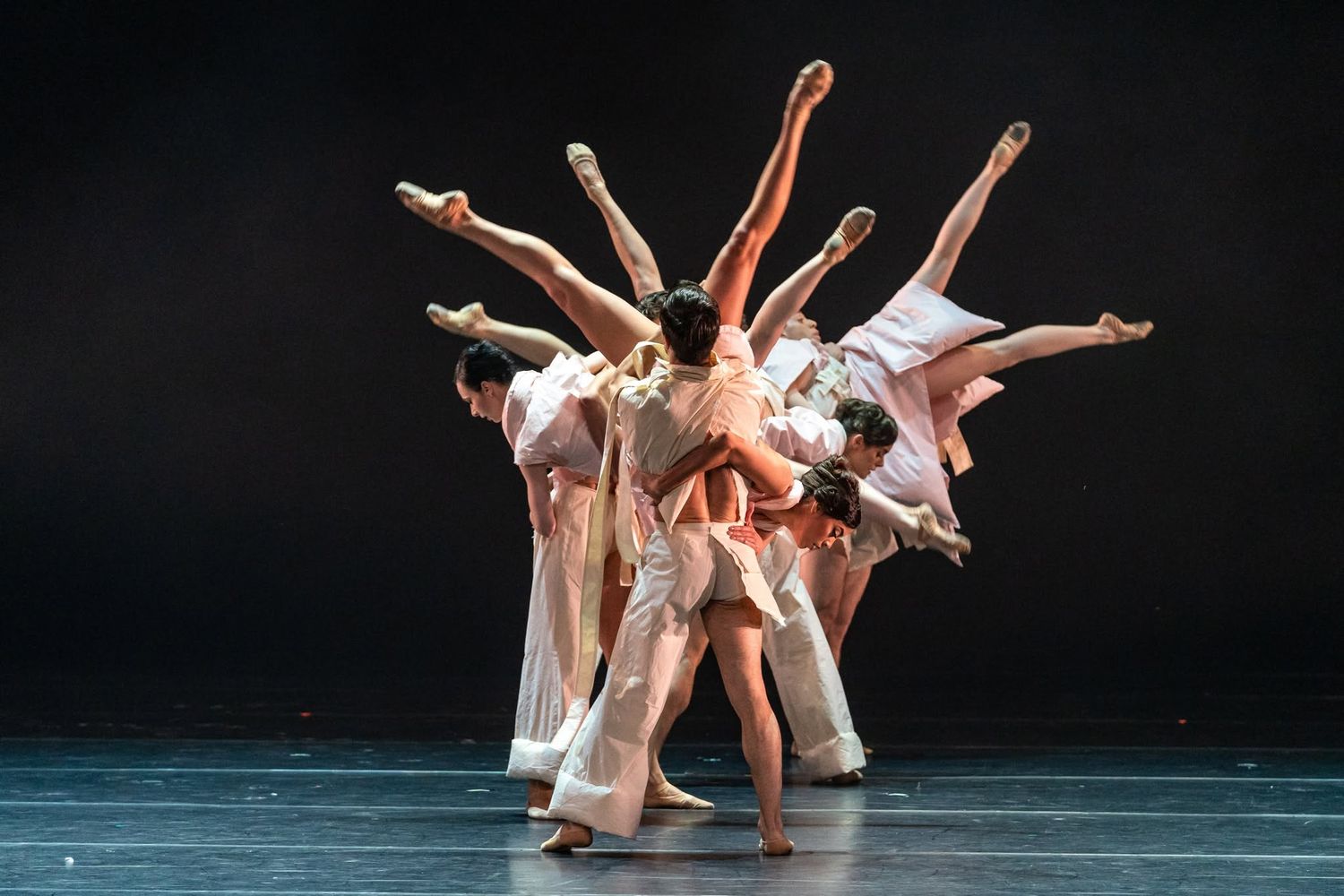 A group of contemporary ballet dancers form a dynamic cluster with extended legs in neutral-toned costumes.