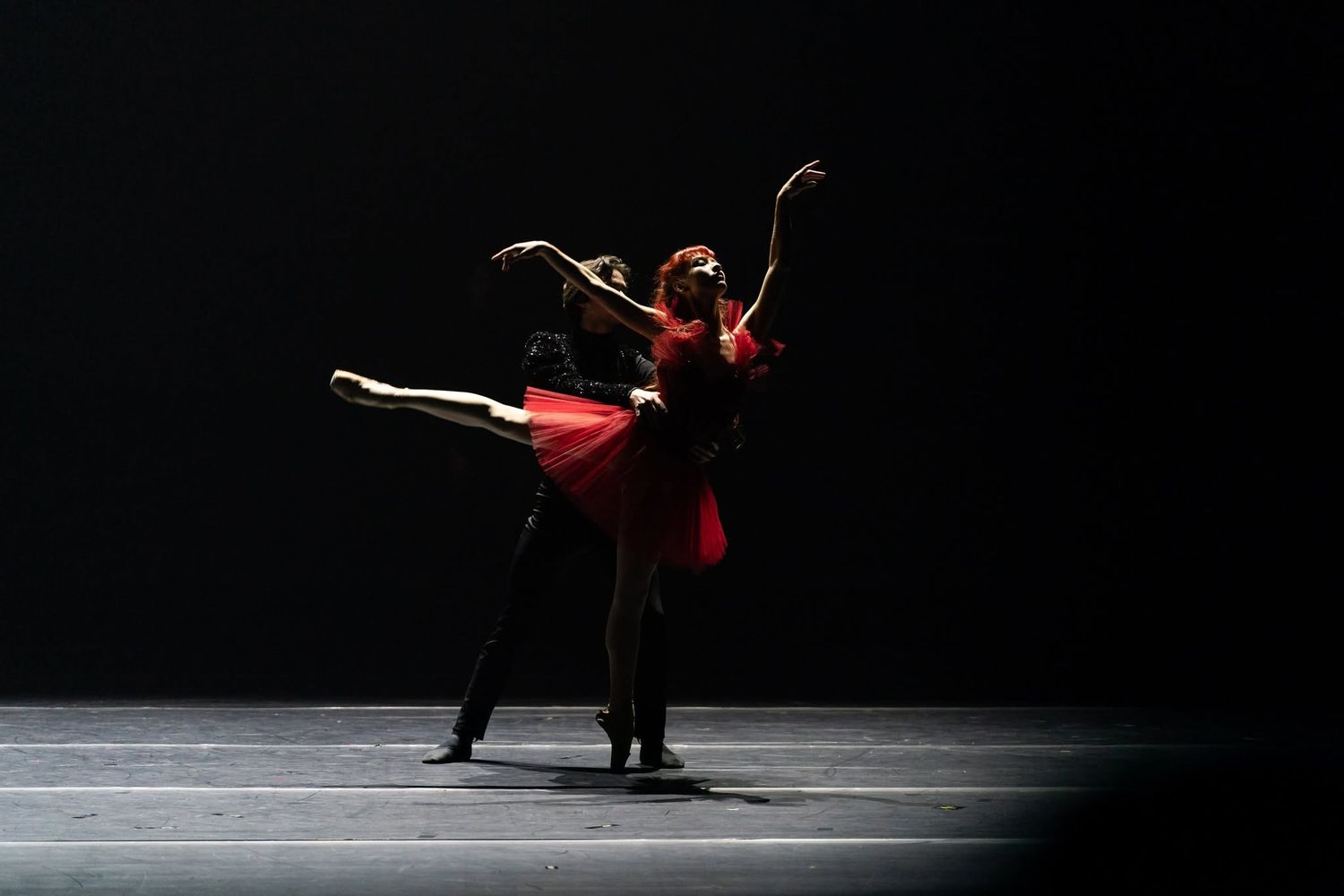 Two ballet dancers perform a dramatic pas de deux in red and black costumes under a dark spotlight.