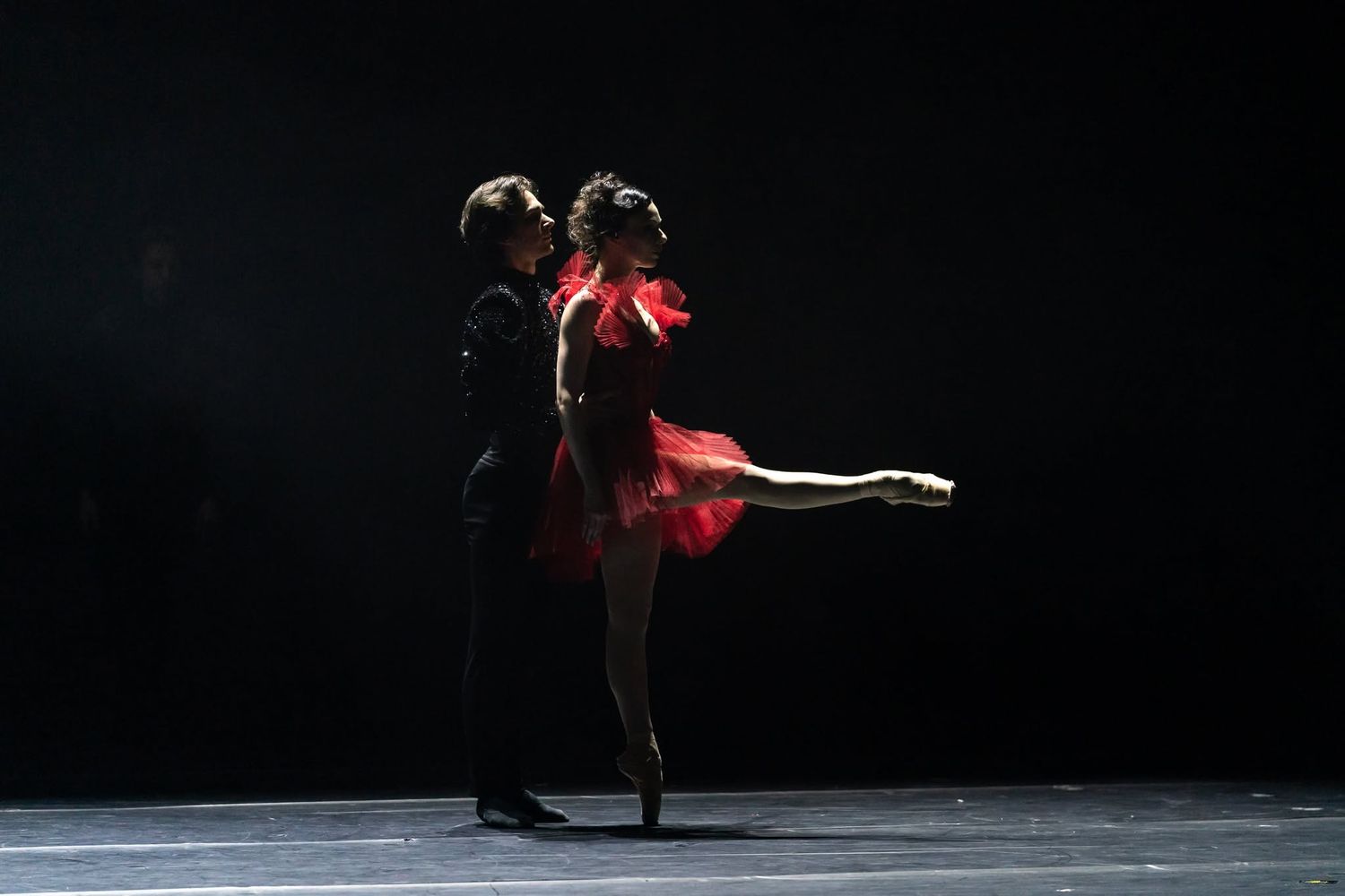 A male and female ballet dancer share an intimate pose in red and black costumes under dramatic dark lighting.