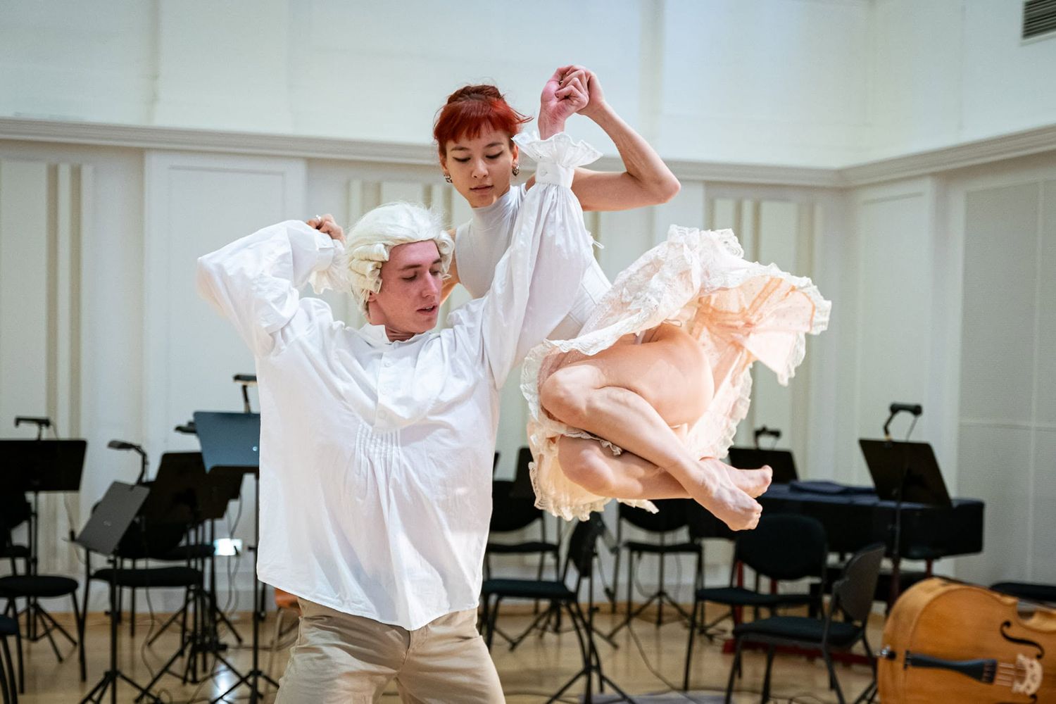 Male dancer lifts female ballet dancer in white costume during orchestra rehearsal room practice session.