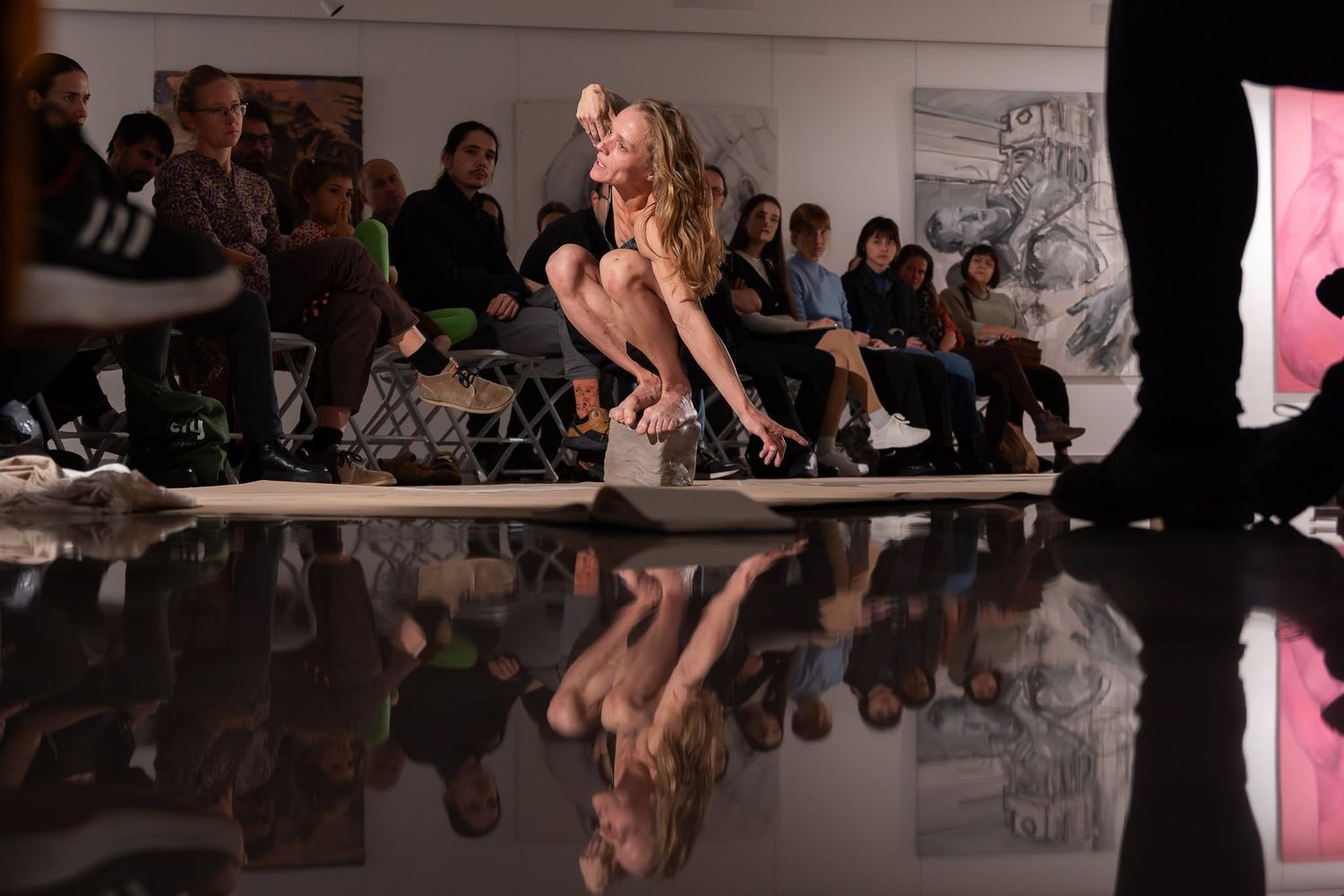 Female dancer performs gracefully on a reflective floor surface before an audience in an art gallery setting.