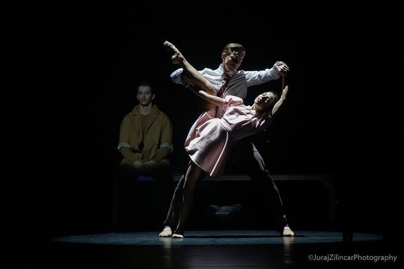 Male dancer lifts female dancer in a pink skirt on a darkened stage during a ballet performance.