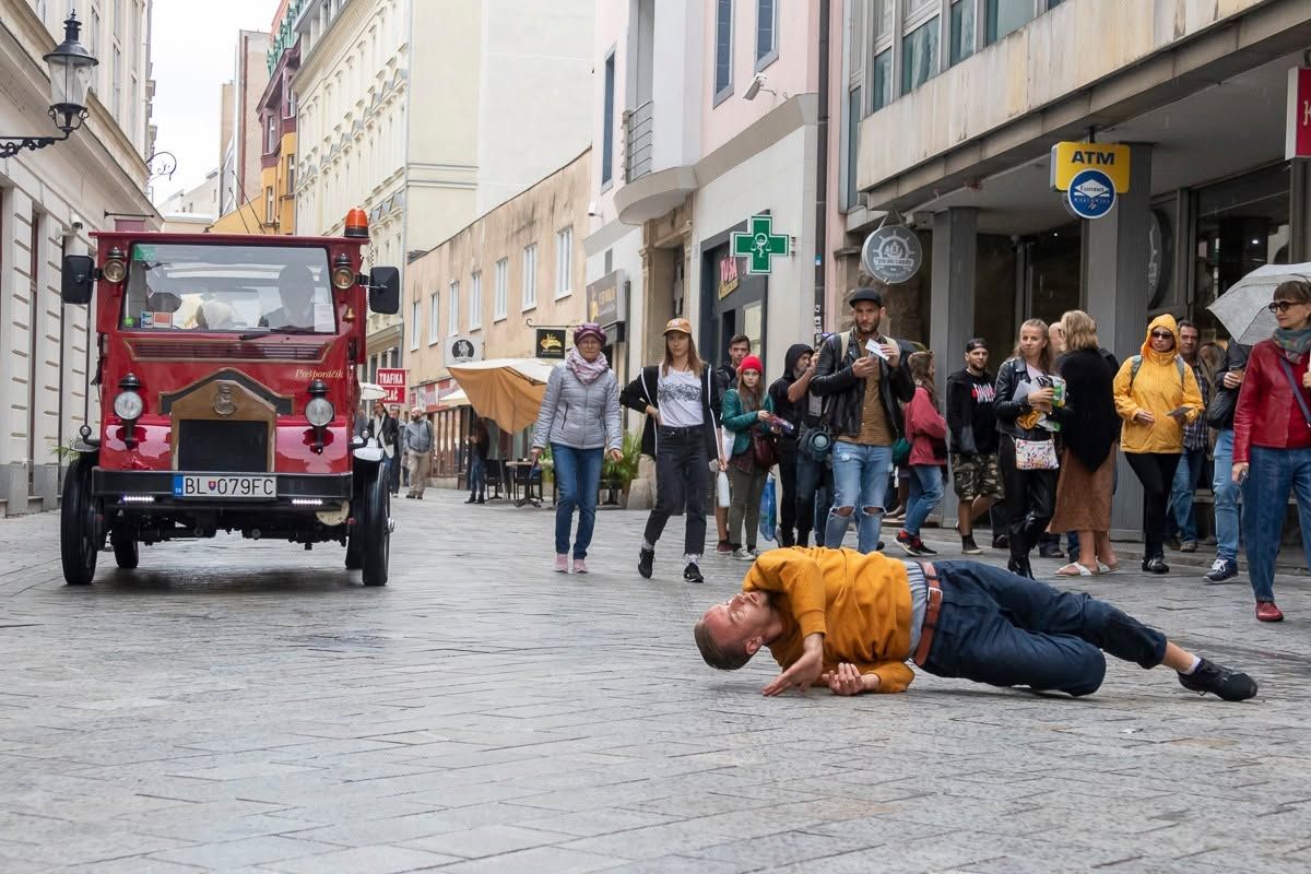 Man lying on cobblestone street in front of red vintage truck with crowd of onlookers in European city.