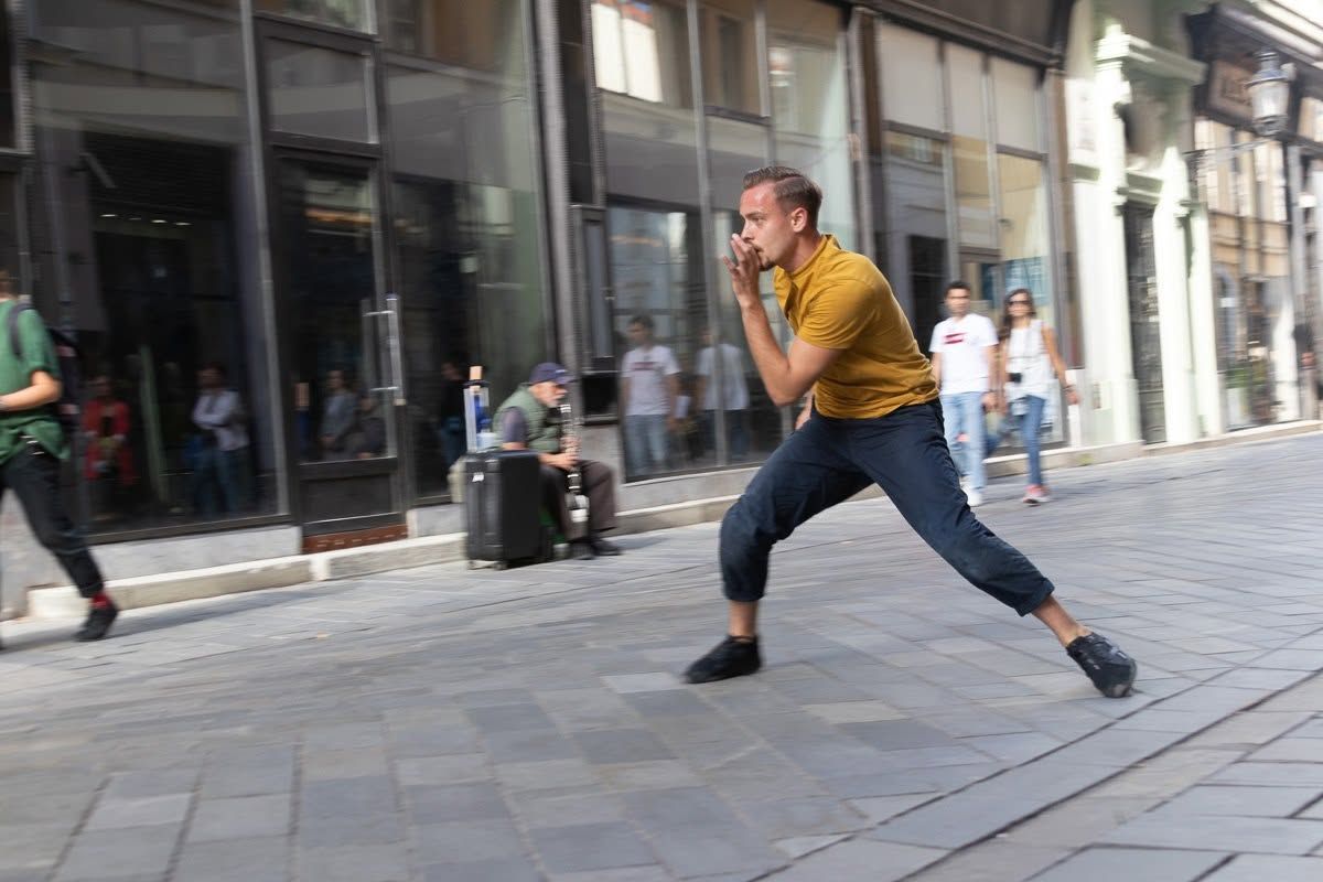 A dancer in a yellow shirt performs a dynamic leap on a busy urban pedestrian street.
