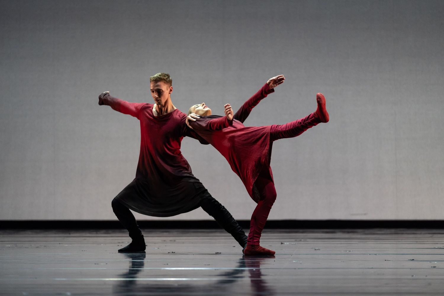 Two contemporary dancers in red and black costumes perform a dynamic partnered routine on a grey stage.