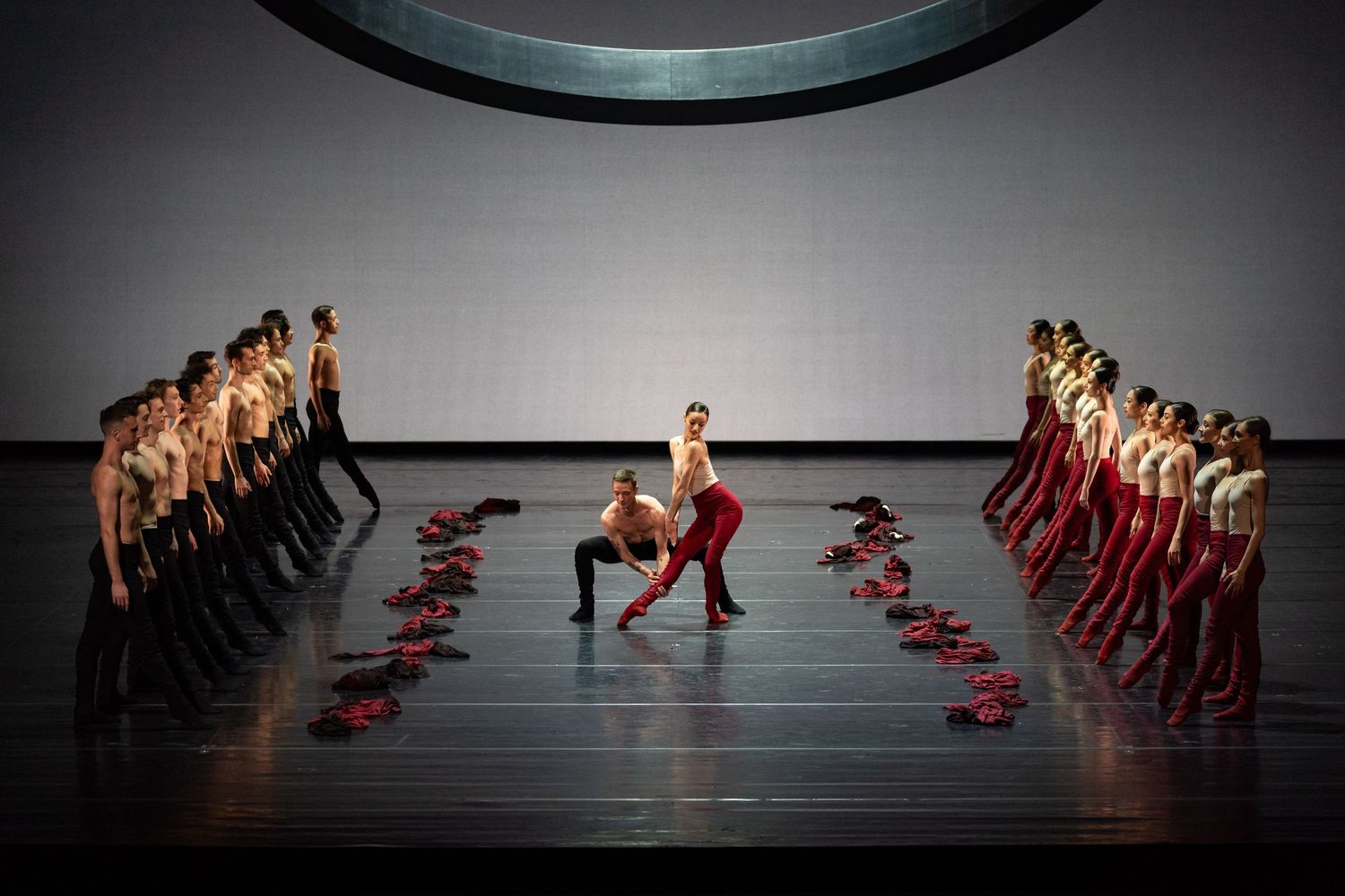 Large ensemble of dancers in red and black flank two soloists in a symmetrical formation on a grey stage.