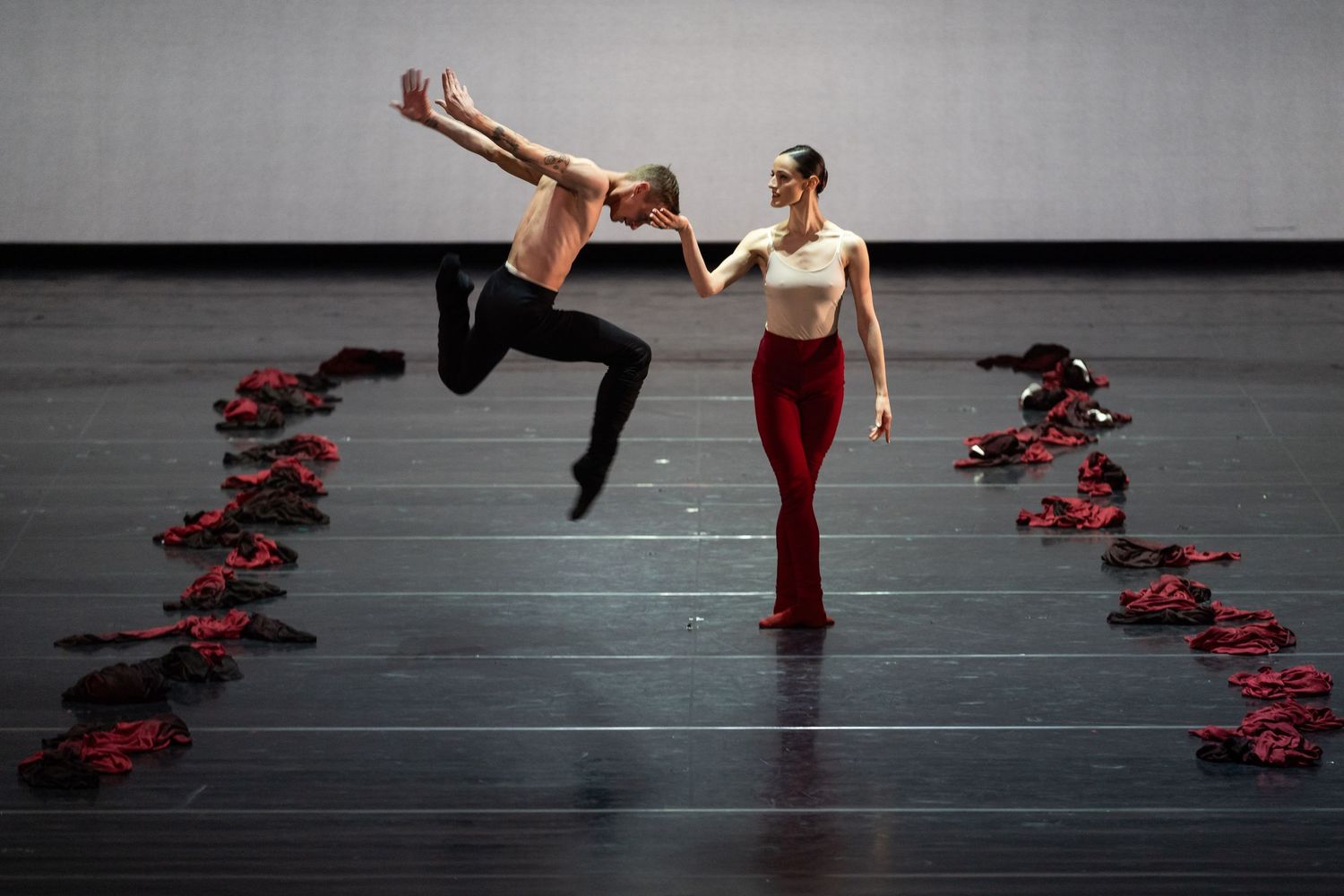 Male dancer leaps beside female dancer in red and white as red garments line the grey stage floor.