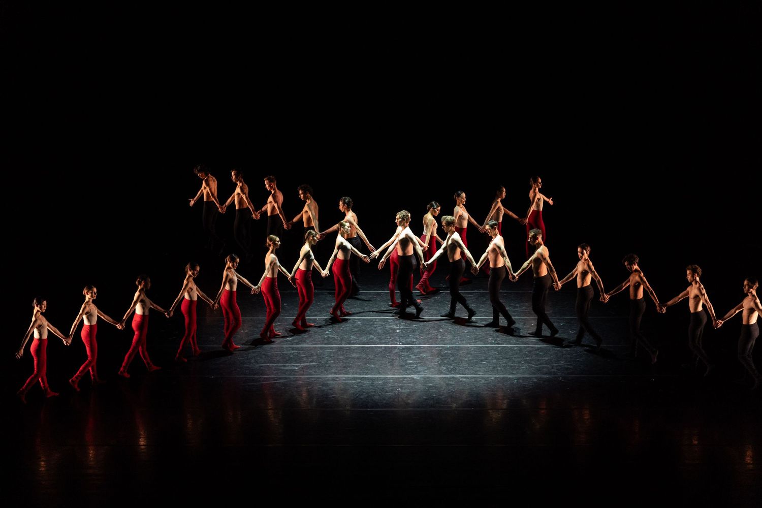 Large ensemble of ballet dancers in red and white costumes performing a dramatic formation on a darkened stage.