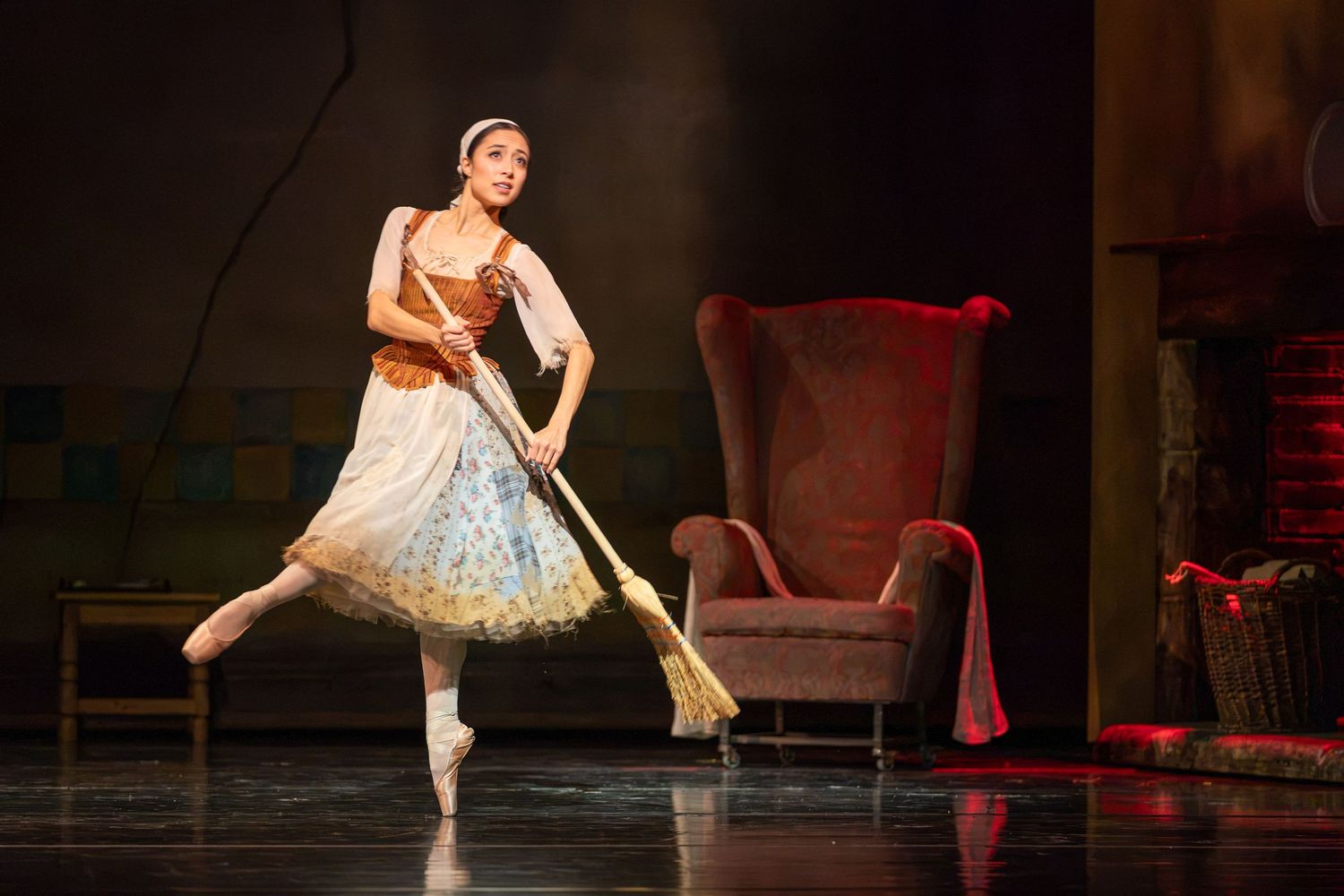 Ballet dancer in costume performs on stage, gracefully balancing on pointe while holding a broom near a red armchair.