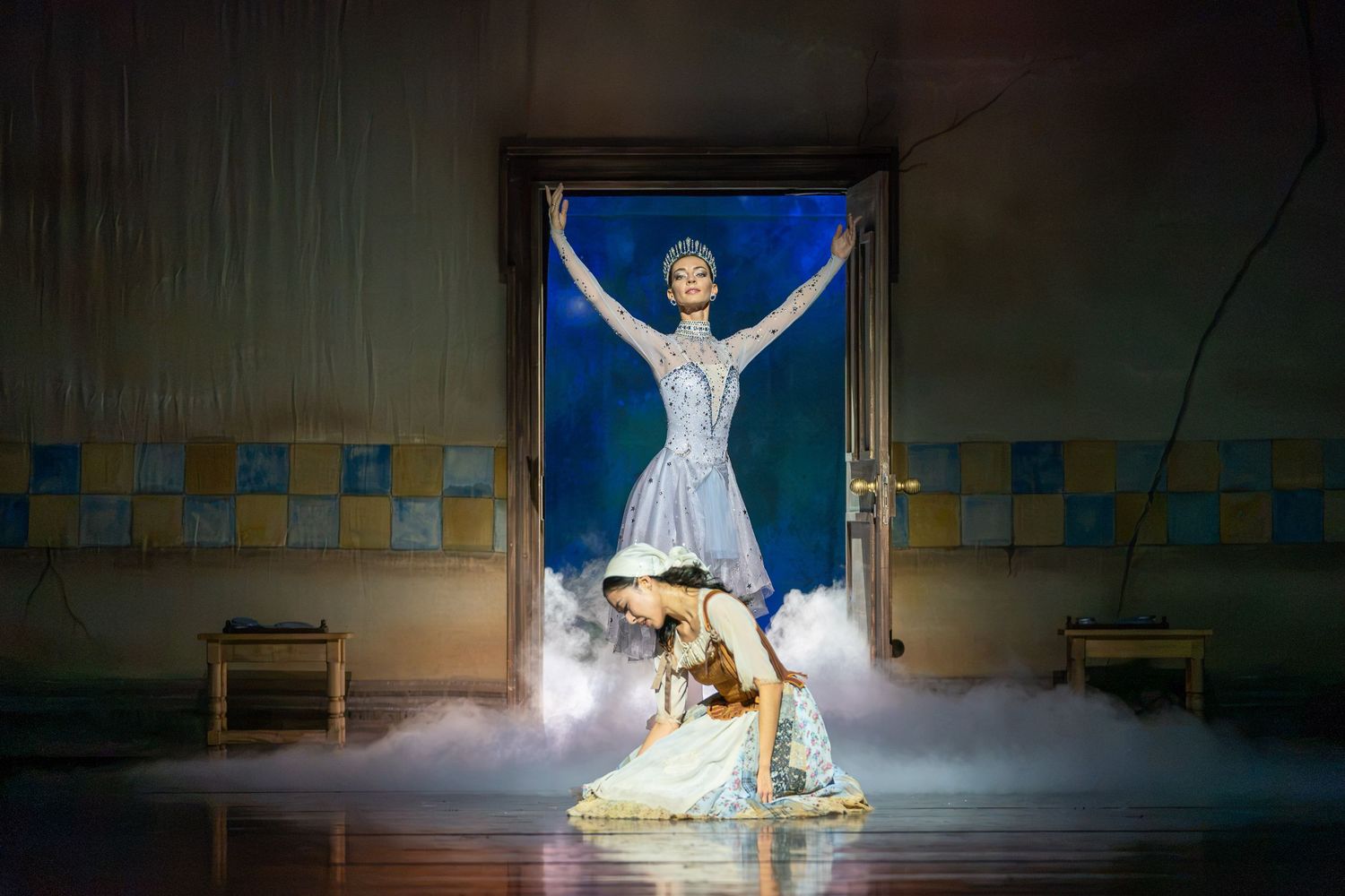 A theatrical ballet performance with a dancer kneeling in mist before a glowing doorway where a ballerina poses triumphantly.