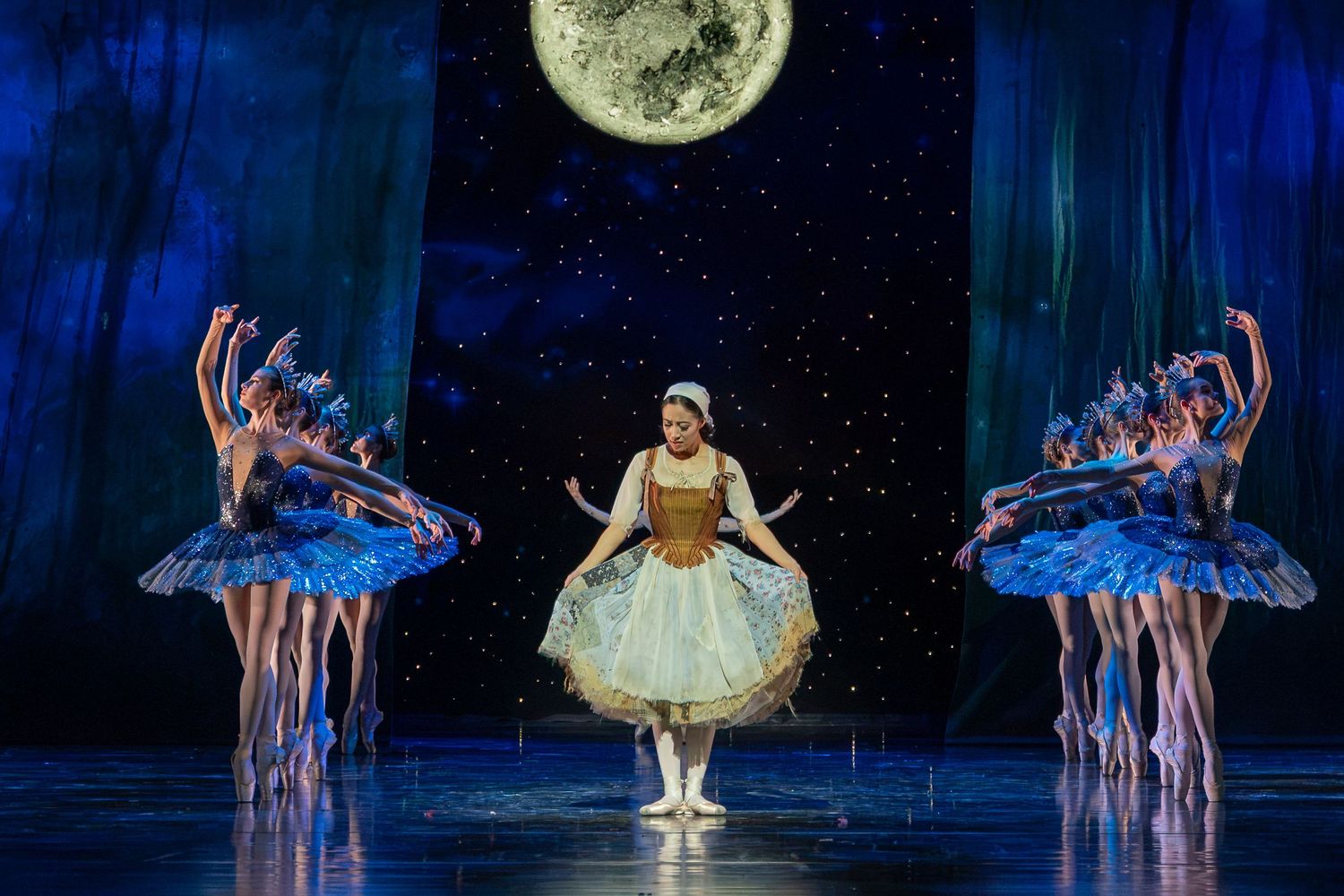 Ballet dancers in blue tutus perform on stage under a large moon backdrop with a central dancer in a gold costume.