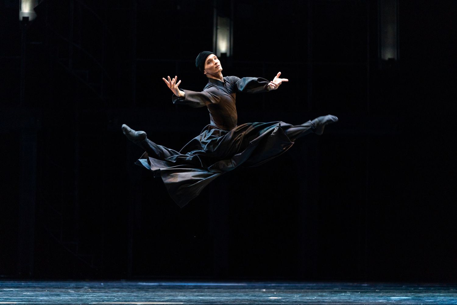 Ballet dancer leaping gracefully in black costume, arms outstretched, captured mid-air on a dark stage.