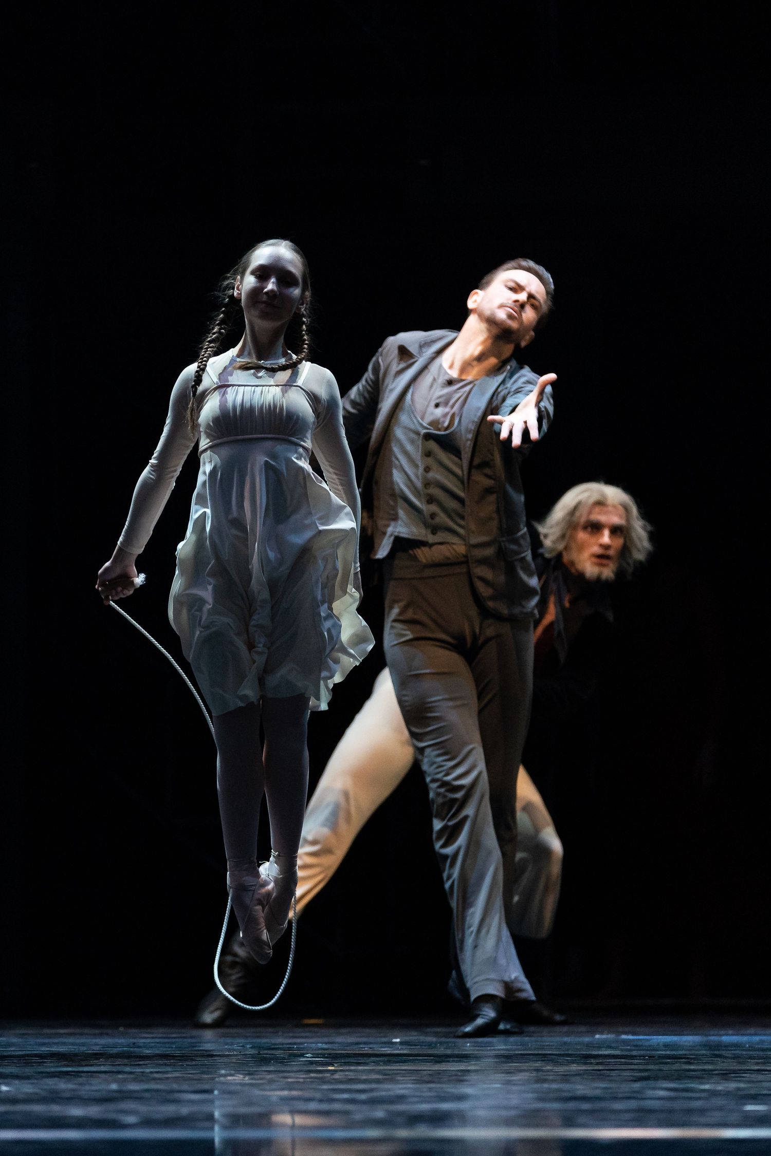 Two contemporary dancers leap dramatically on dark stage with mysterious figure lurking in the background.