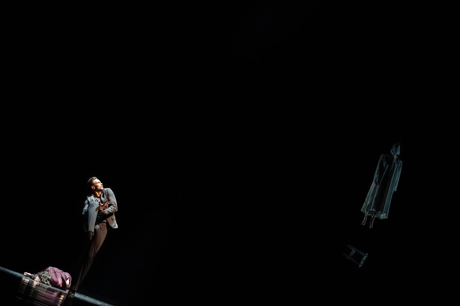 Two performers on a dark stage, one illuminated in spotlight with luggage, another ghostly figure visible in the distance.