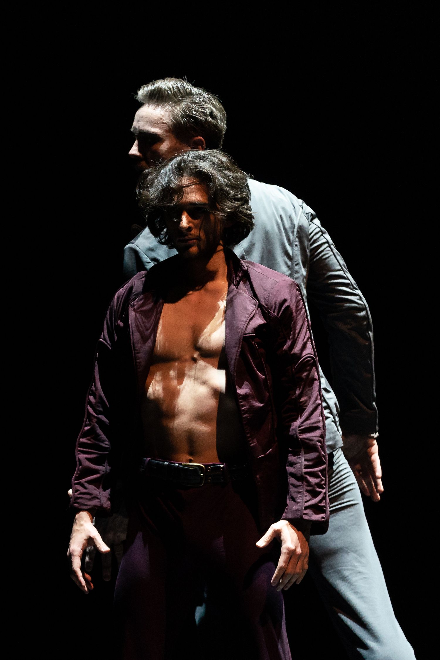 Two male dancers in dramatic pose, one shirtless in open purple shirt, the other in grey, against black background.