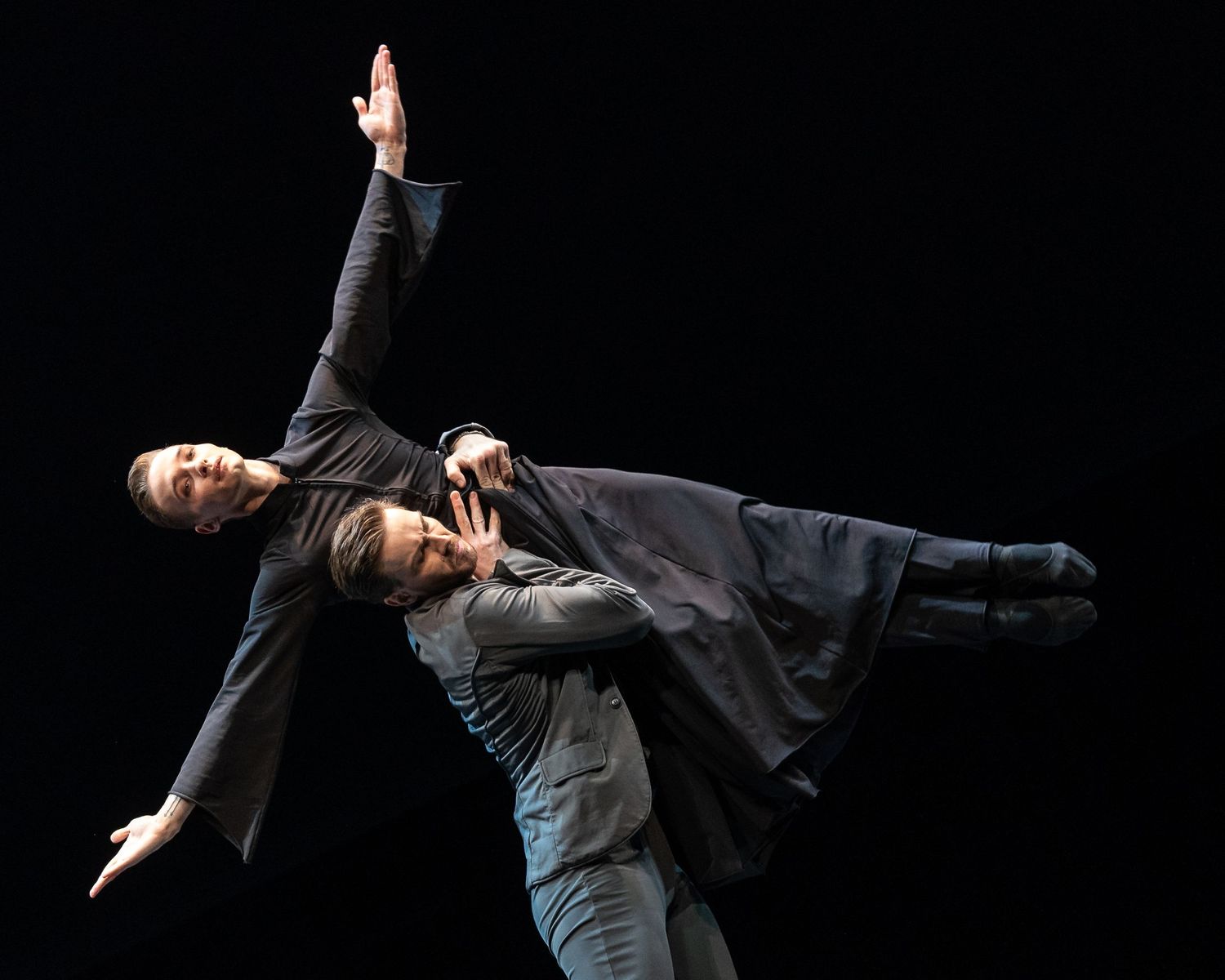 Two contemporary dancers performing a dramatic lift, one extended horizontally in flowing black costume on dark stage.