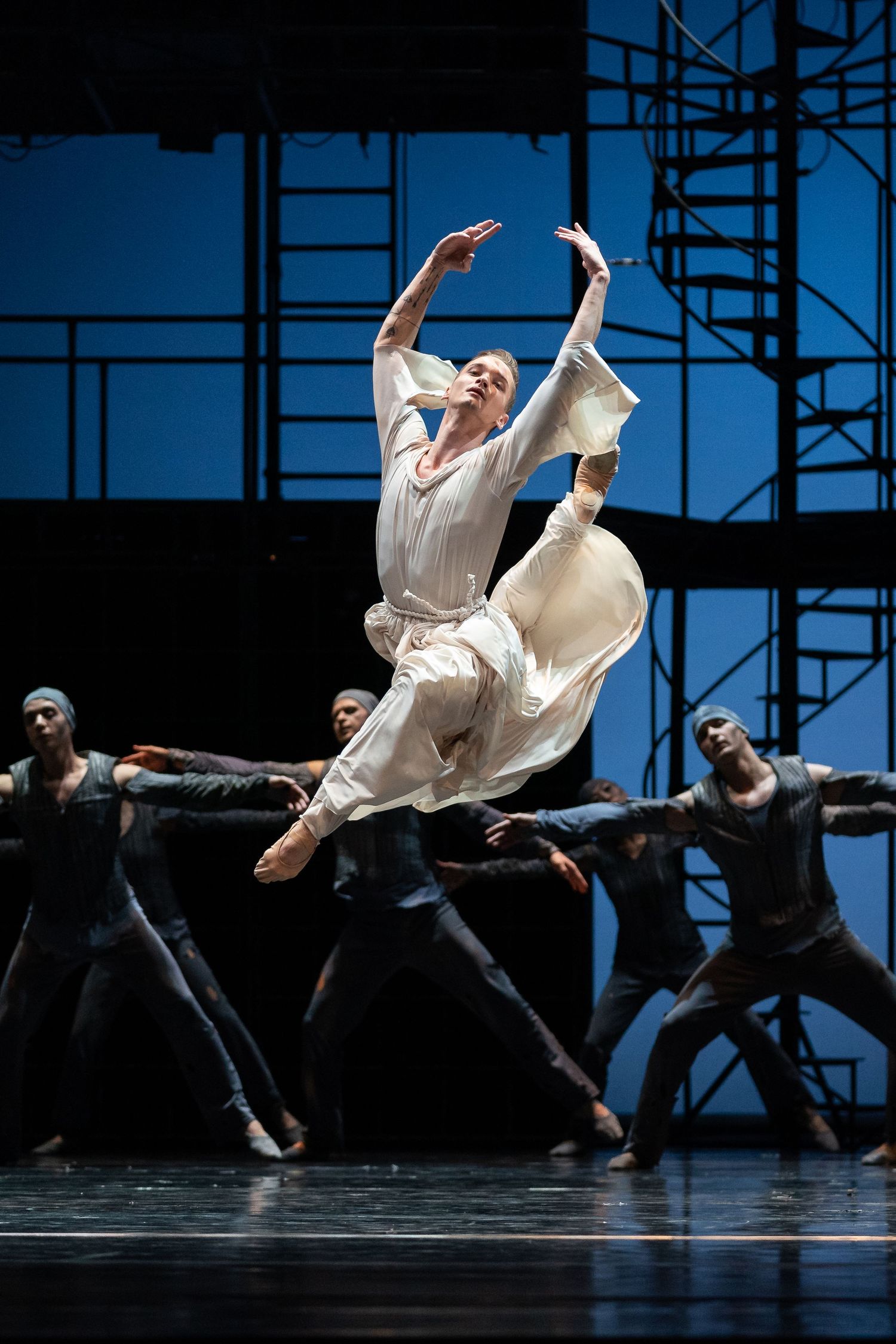 Female ballet dancer leaps in white costume while ensemble in black poses on a blue-lit industrial stage set.