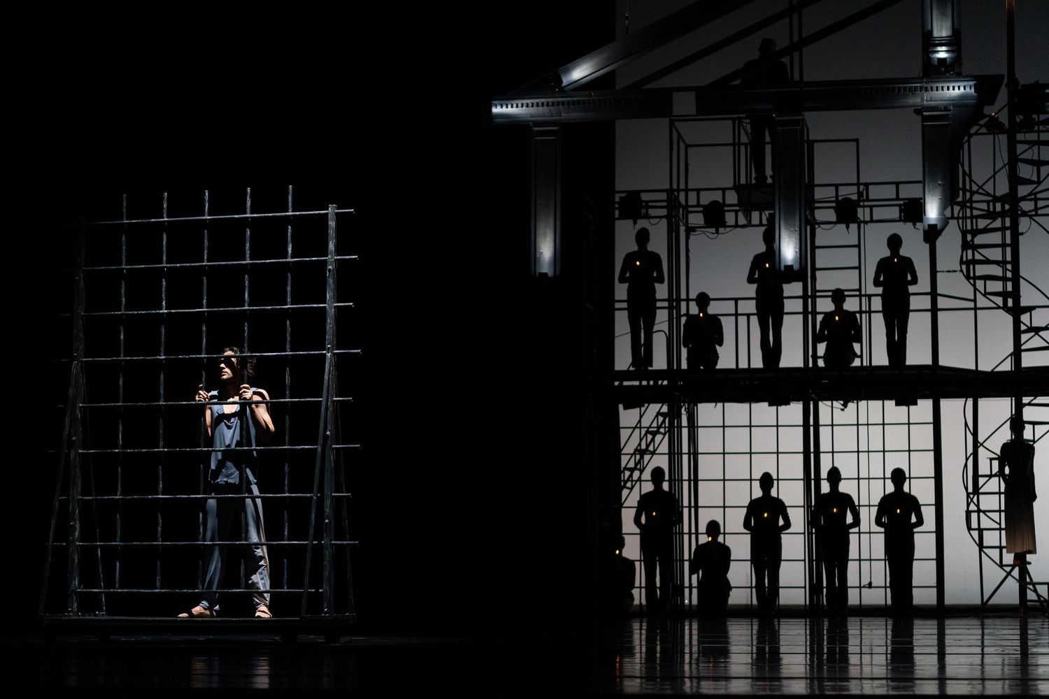 Lone dancer stands by metal grid structure as silhouetted figures line scaffolding in dramatic theatrical lighting.