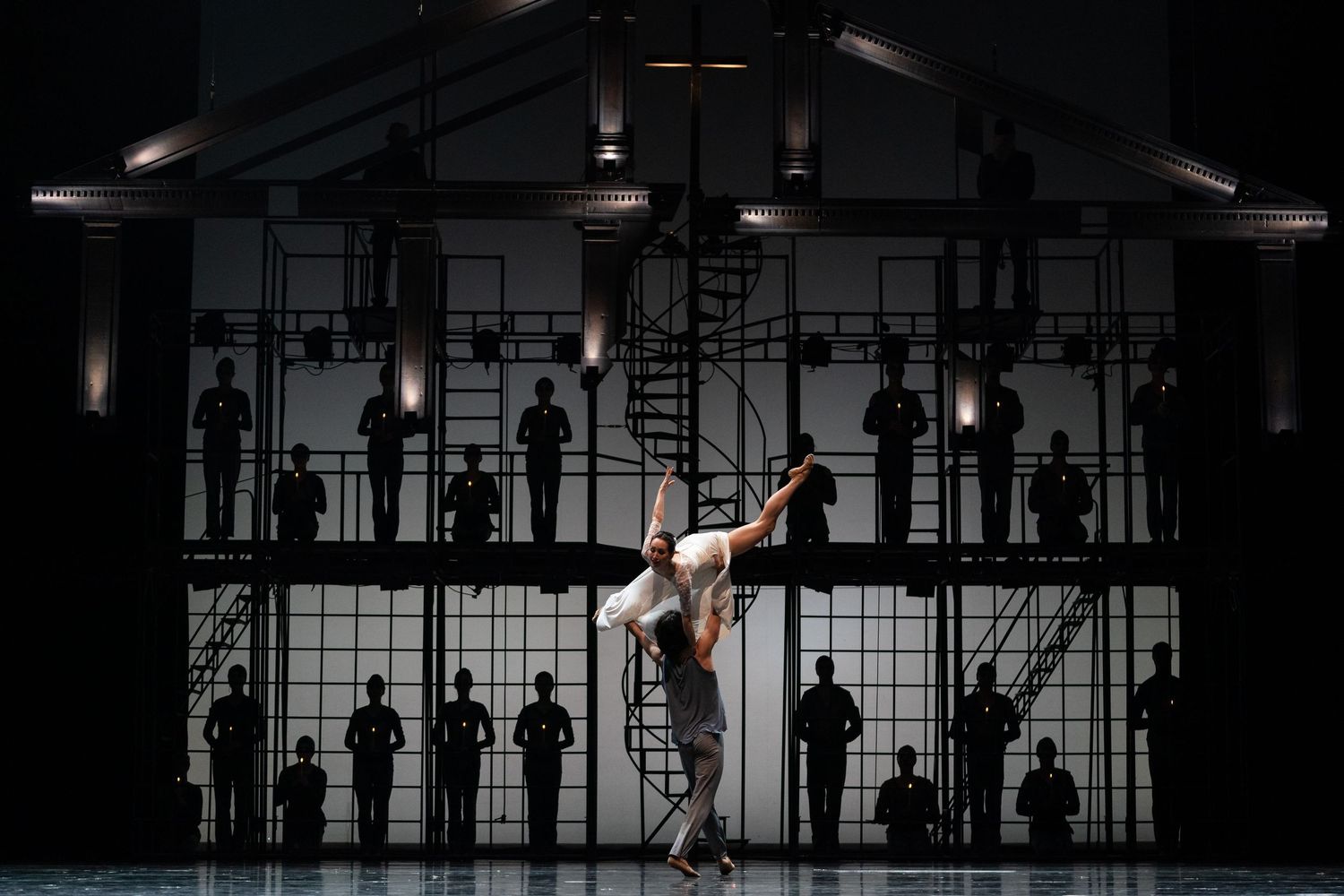 Two dancers perform a dramatic lift before a silhouetted ensemble on scaffolding in a striking stage production.