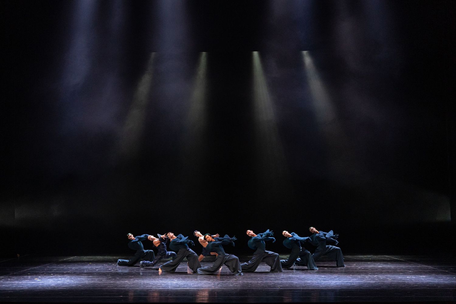 Group of dancers in blue costumes perform synchronized floor choreography under dramatic spotlights on stage.