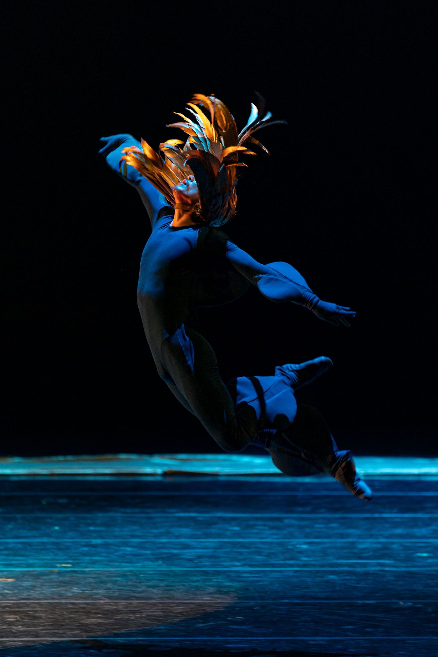 Dancer leaps dramatically wearing colorful feathered headdress under blue and orange stage lighting.