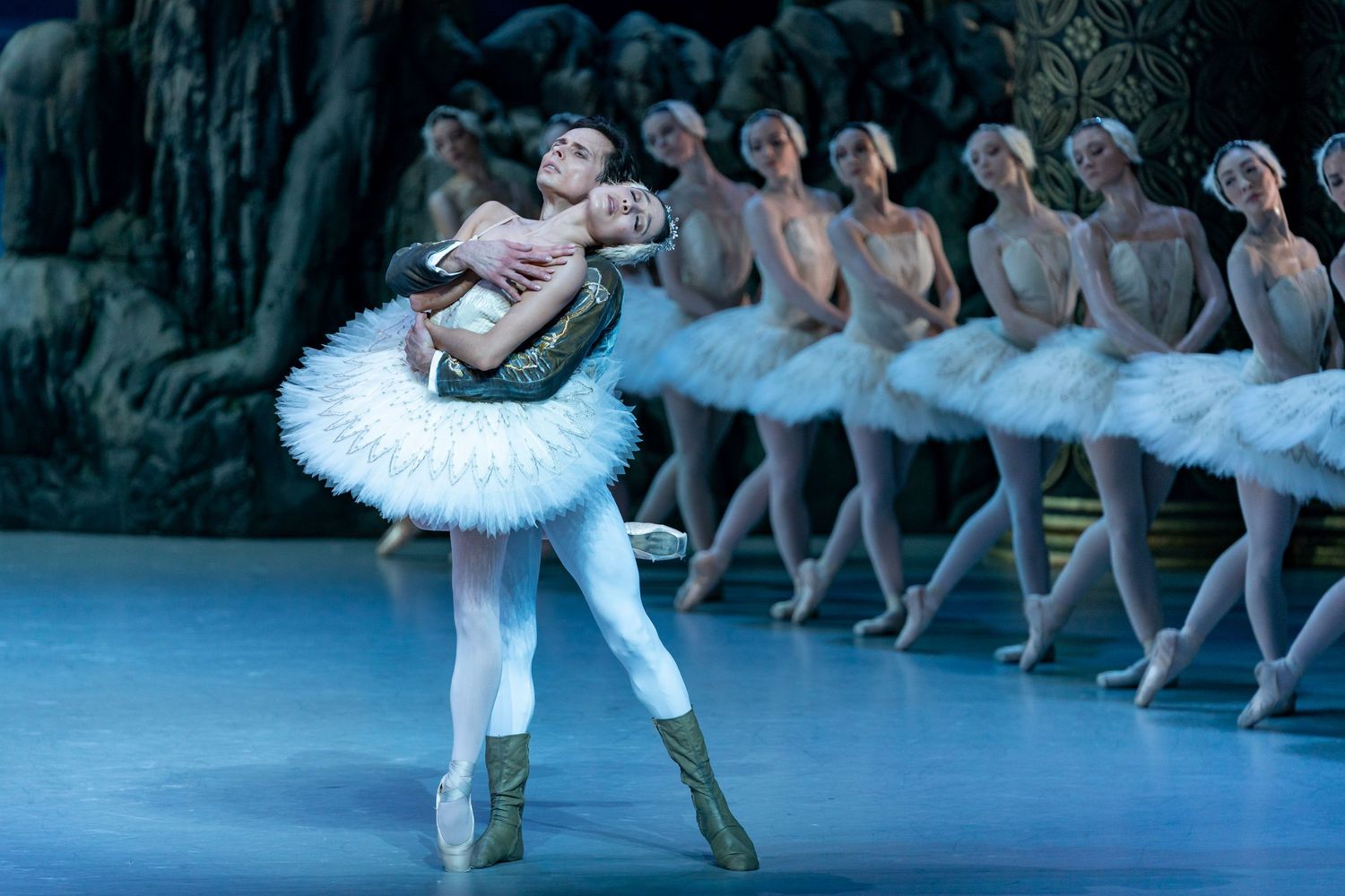 Ballerina in white tutu is lifted by partner during Swan Lake performance with corps de ballet in background.
