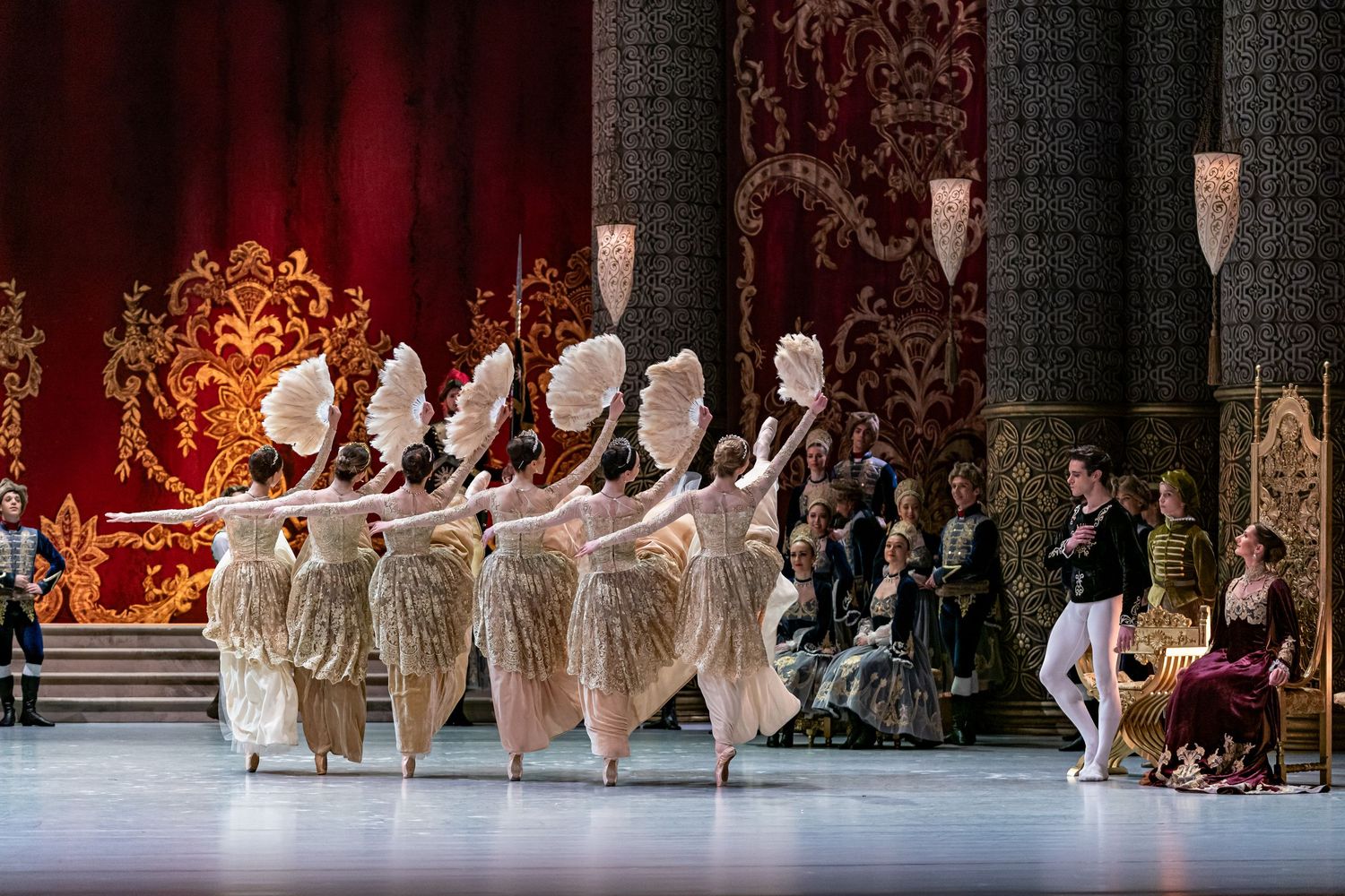 Ballet dancers in feathered white costumes perform before seated court audience on ornate red and gold stage.