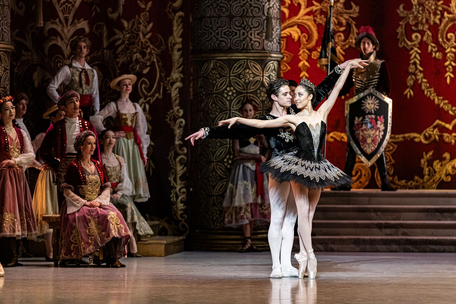 Ballerina in black tutu performs with male partner before royal court audience on richly decorated red stage.