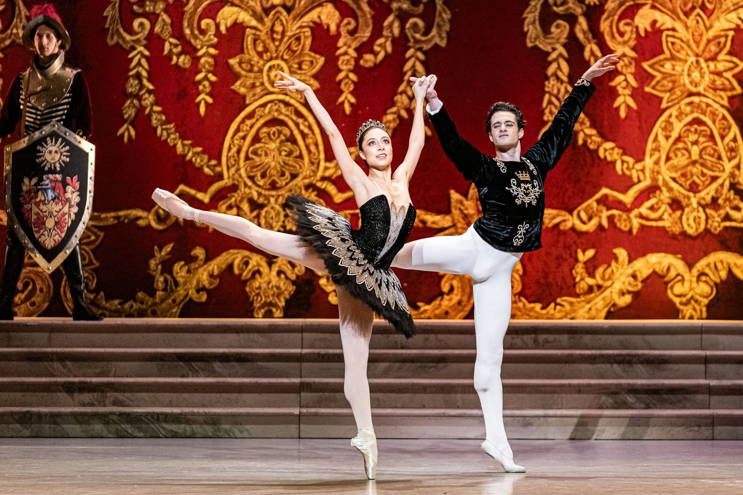 Ballerina and male dancer perform dramatic pas de deux in black and white costumes against ornate red backdrop.