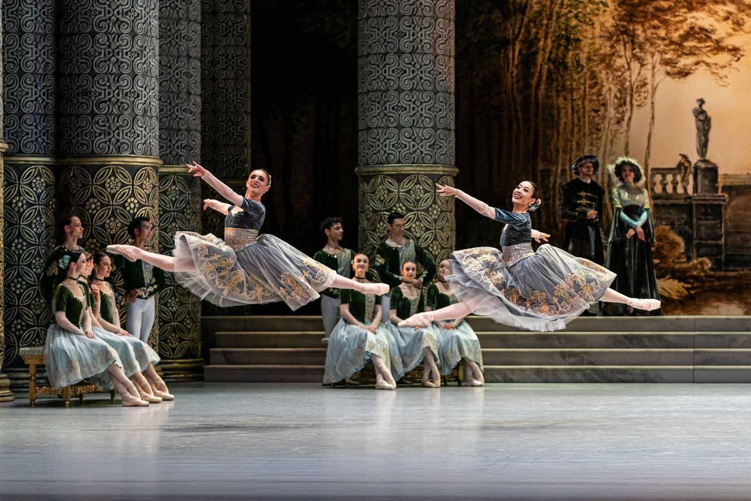Three female ballet dancers leap gracefully in floral costumes on an ornate classical stage set with columns.