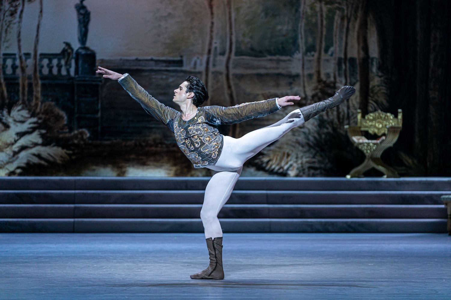 Male ballet dancer performs elegant arabesque in ornate costume on classical stage set during ballet performance.
