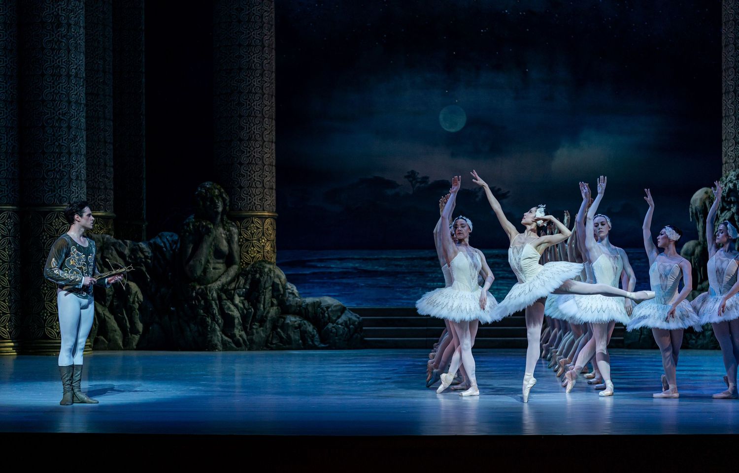 Male ballet dancer watches as ballerina performs with corps de ballet in white tutus on dramatic blue-lit stage.