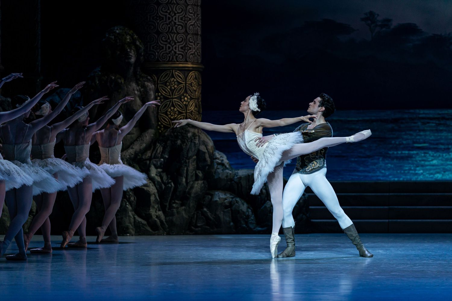Male and female ballet dancers perform elegant pas de deux in Swan Lake with corps de ballet in background.