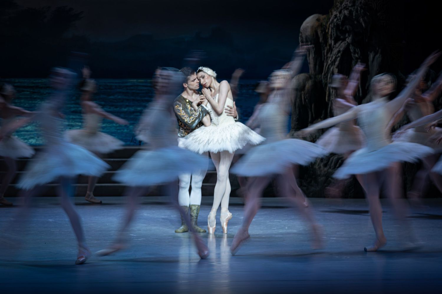 Two ballet dancers embrace center stage surrounded by blurred corps de ballet in white tutus during Swan Lake performance.
