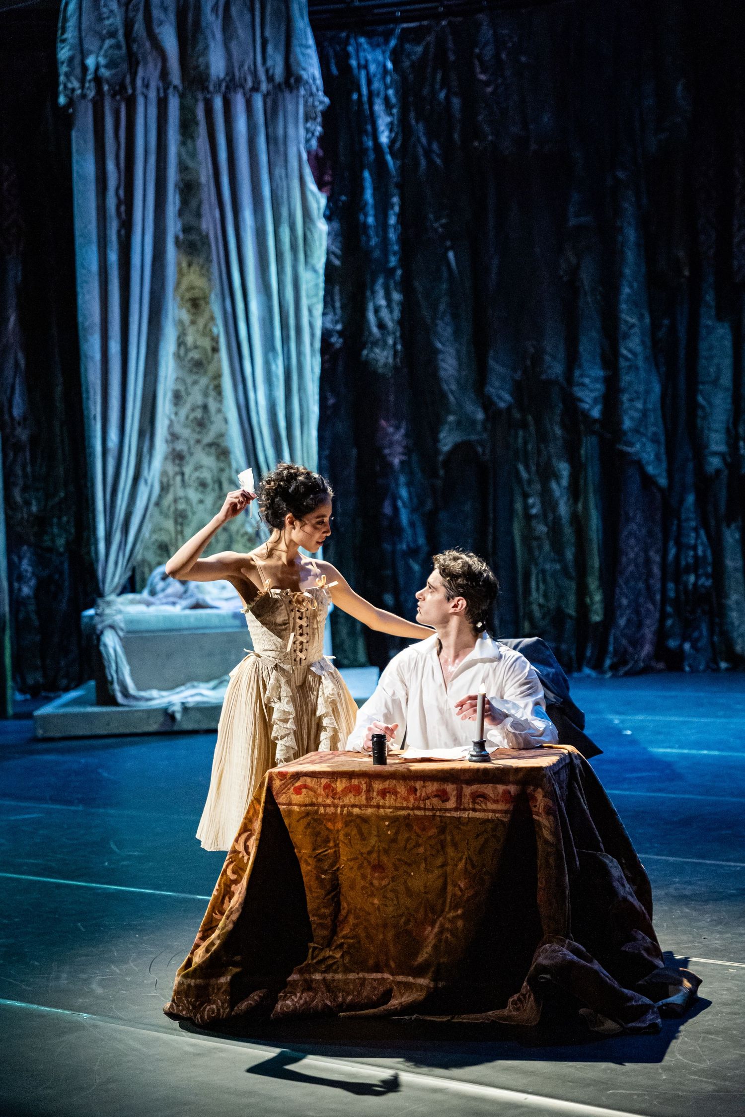 A ballet dancer in a period gown stands beside a seated man at an antique desk in a dramatic stage scene.