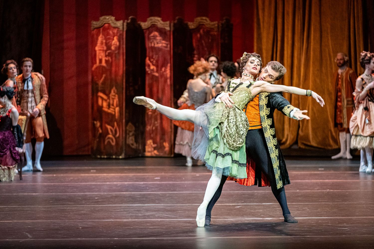 Two ballet dancers perform an elegant pas de deux in ornate period costumes on a grand theatrical stage.