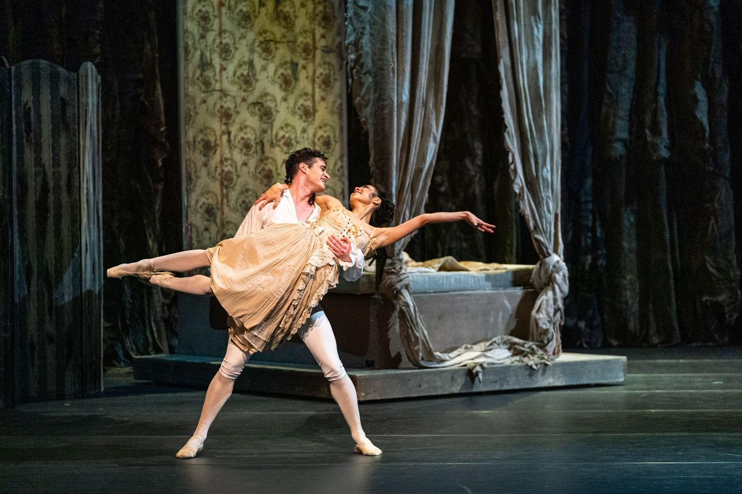 Male and female ballet dancers perform classical pas de deux in ornate period costumes on decorated stage.
