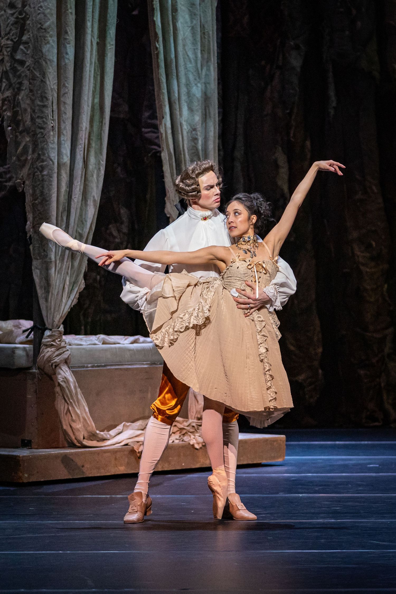 Ballet couple performs classical pas de deux in elegant period costumes against dramatic draped backdrop.