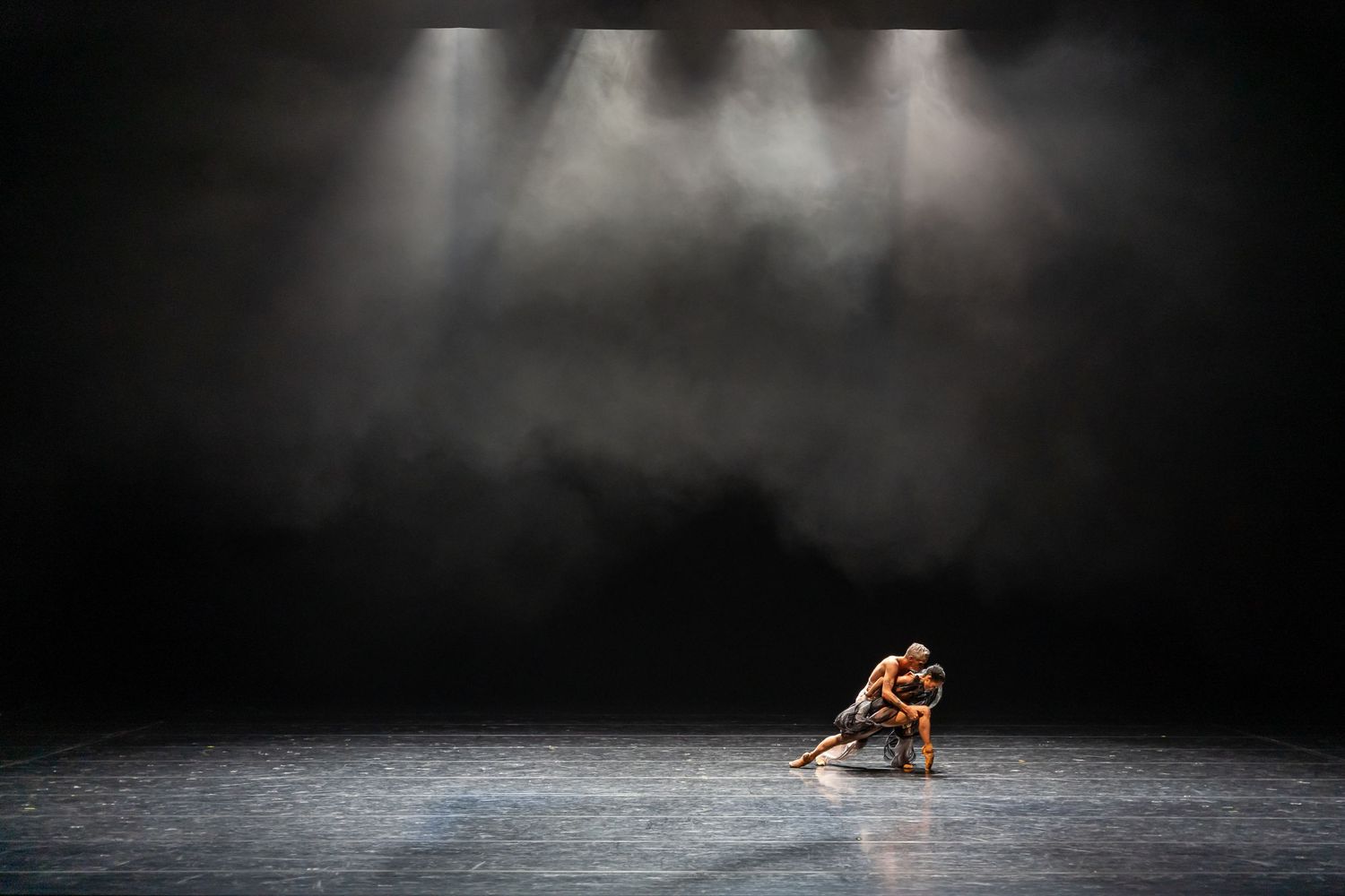 Two dancers share an intimate contemporary dance moment in a vast, misty, dramatically lit stage.