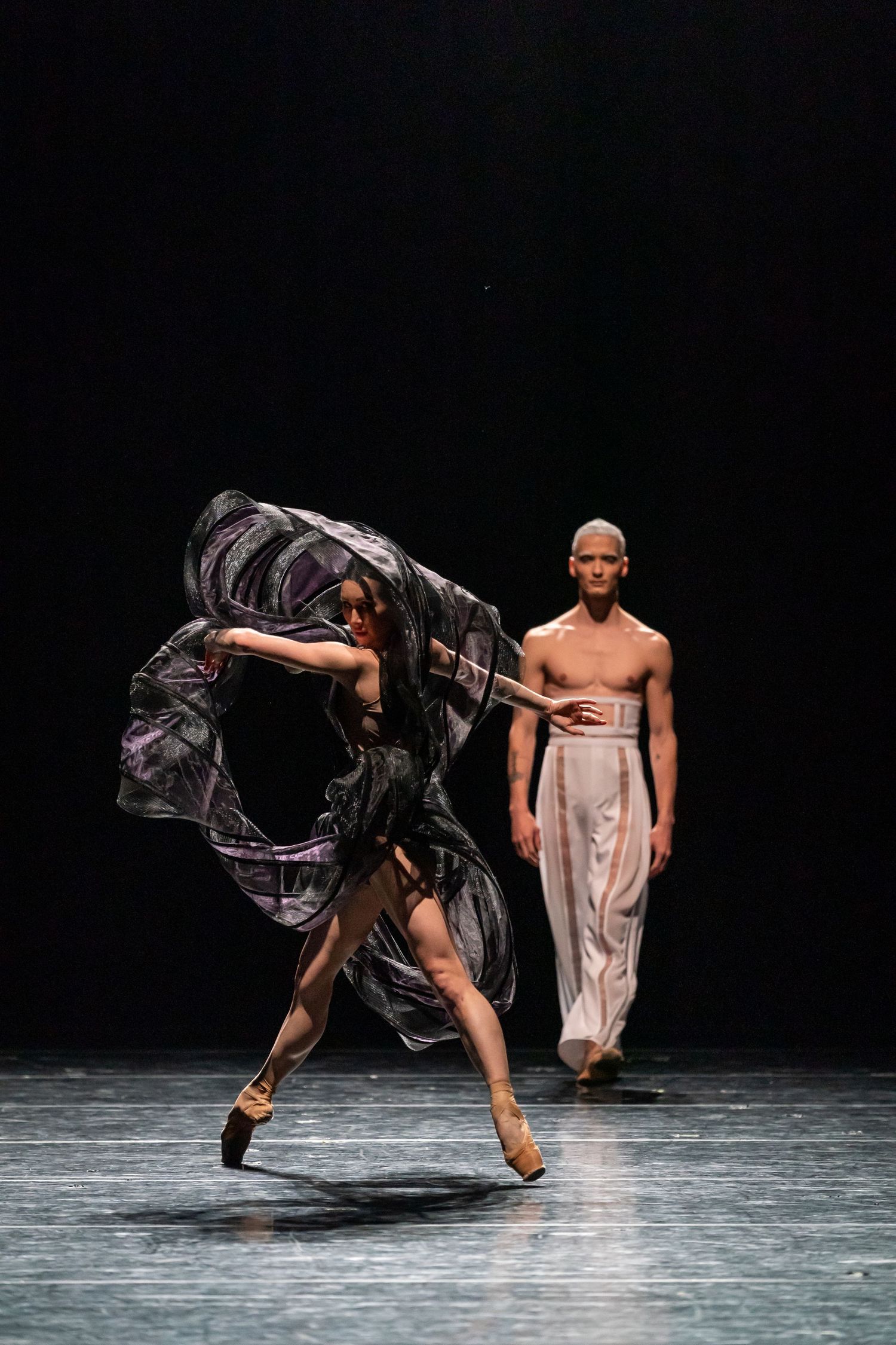 Female ballet dancer in flowing black dress performs on pointe while male dancer walks behind her.