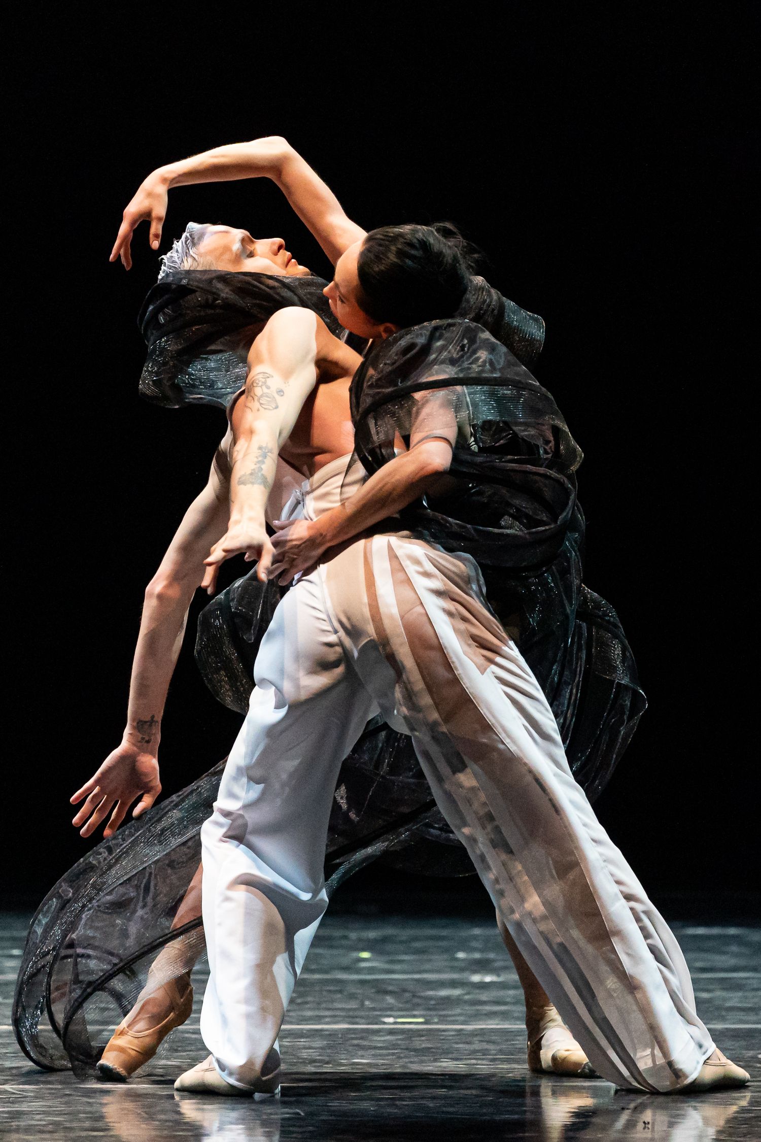 Two ballet dancers in an intimate contemporary pas de deux, one in flowing dark costume on stage.