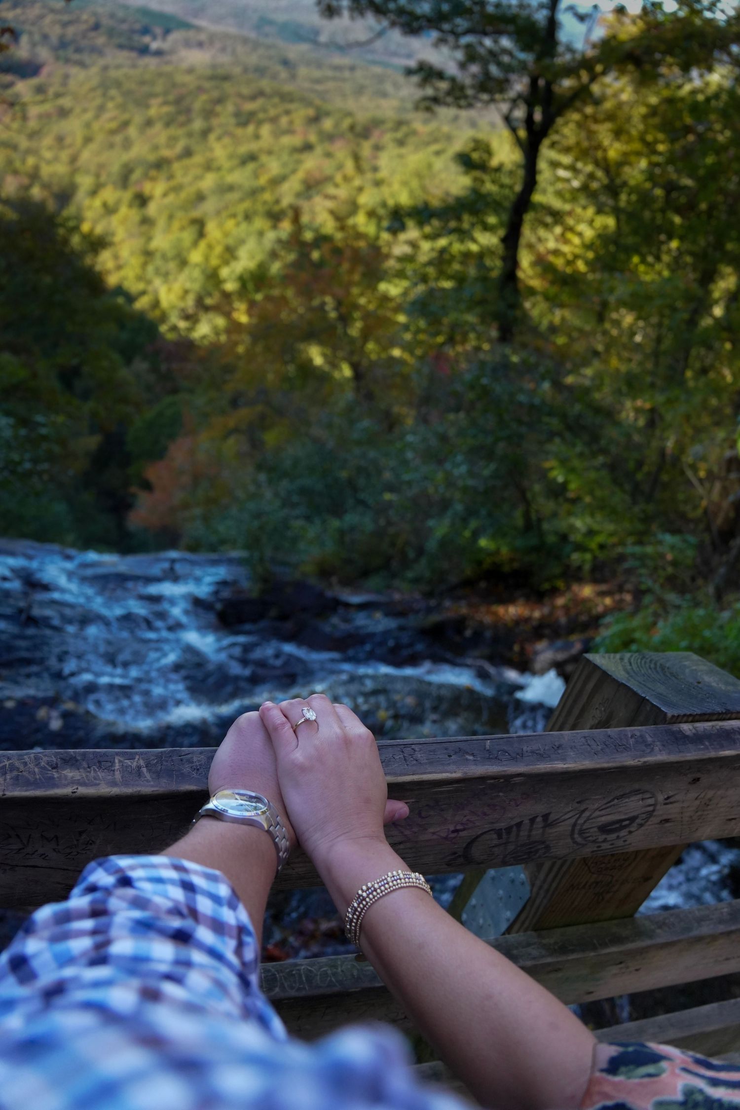 Couple holding hands on a wooden railing overlooking a scenic waterfall and lush green forest valley.