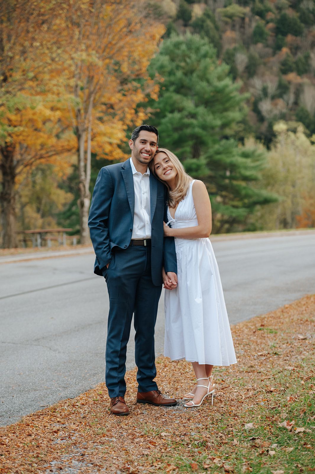 A couple in wedding attire shares intimate moments on a scenic autumn road in upstate NY  lined with colorful fall foliage.